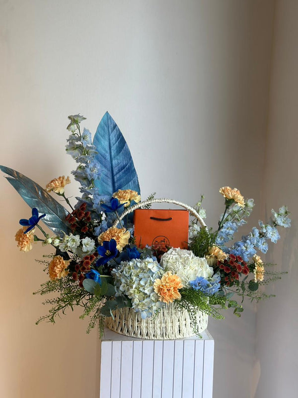 Floral arrangement with a basket containing an orange bag on a white pedestal against a plain background